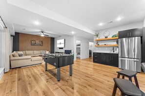 A modern living room with a foosball table, a sofa, and a kitchen area with a refrigerator and cabinets. - Panoramic Table Rock Lake Views with Hot Tub, Games & Free Branson Attraction Tickets (Hollister)