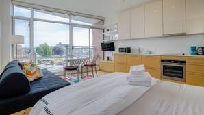 Ein modernes Hotelzimmer mit großem Balkon, einem Bett mit weißer Bettwäsche, einer Sitzecke, einem Esstisch mit Stühlen, einer Küchenzeile inklusive Geräte und Blick auf die Stadt.