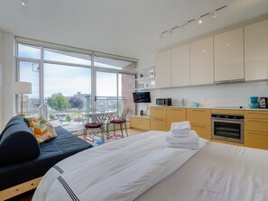 Una moderna camera d'albergo con un ampio balcone, un letto con lenzuola bianche, un divano, un tavolo da pranzo con sedie, angolo cottura completo di elettrodomestici e vista sulla città.