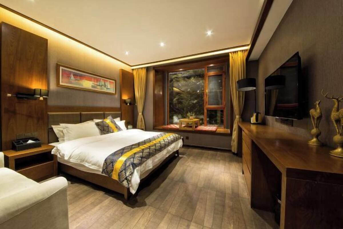 A hotel room with a bed, bedside table, lamp, TV, and window with curtains.