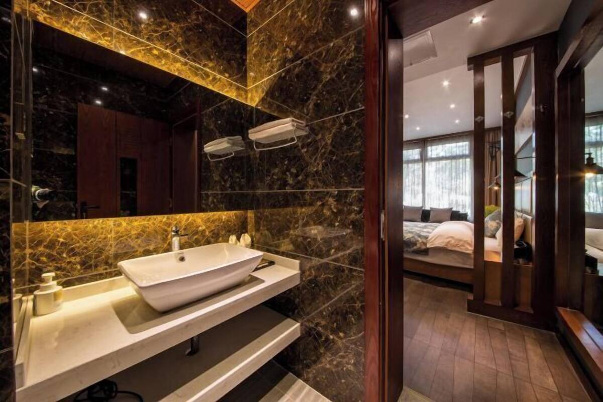A modern bathroom with a large vessel sink, dark marble walls, and a visible bedroom with a bed and window.