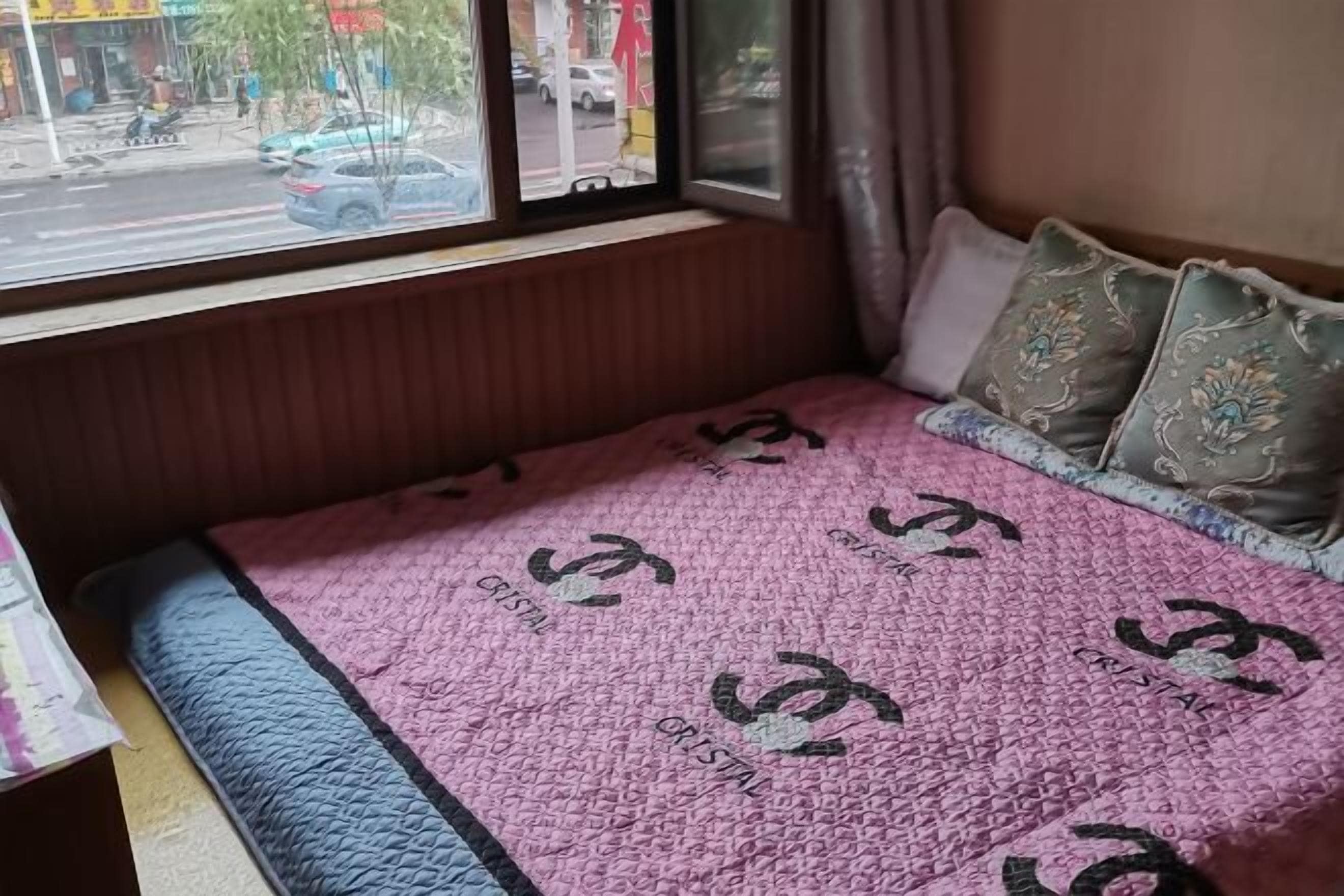 A bed with a pink blanket featuring a repeated logo, a window with a view of the street, and a wooden headboard.