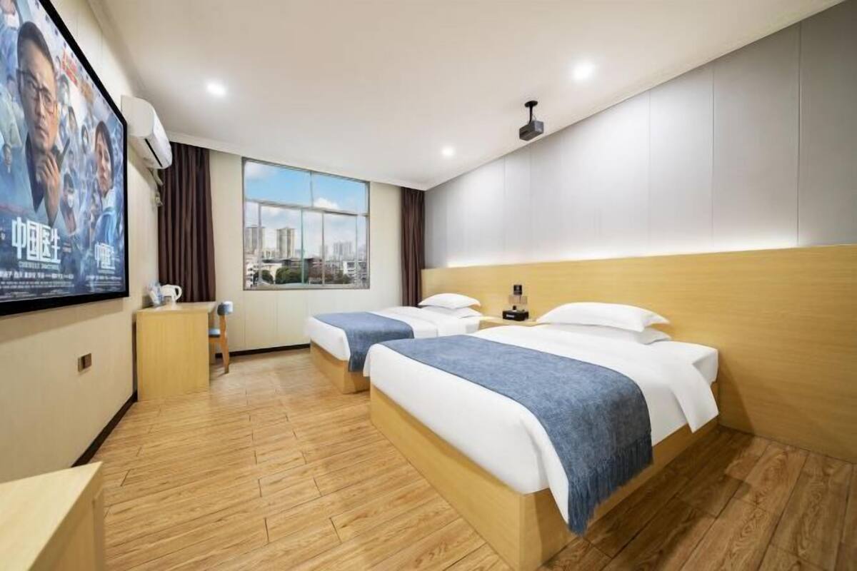 A hotel room with two beds, a large window, a city view, and a poster on the wall.