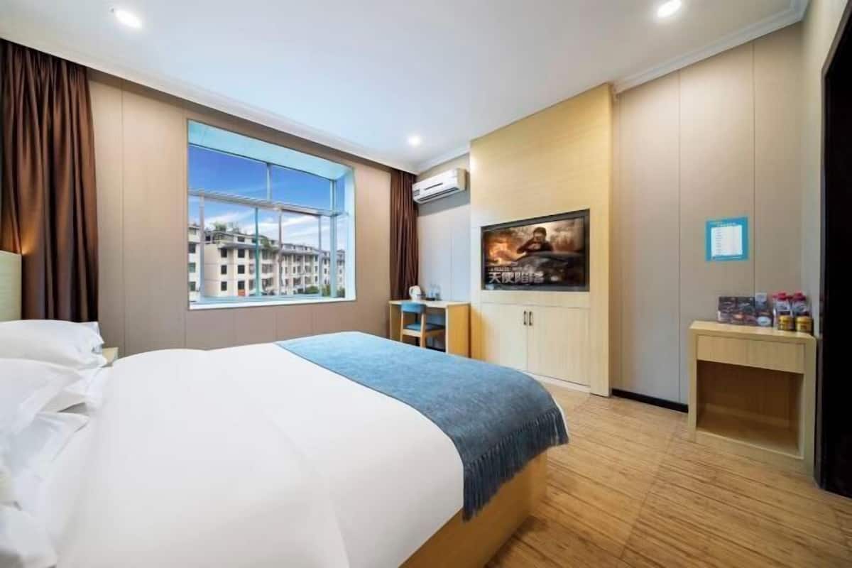 A hotel room with a large bed, a desk, a chair, a window with a city view, and a small table with items on it.