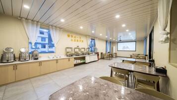 A spacious room with a long table and chairs, a kitchenette area, and a projection screen on the wall.