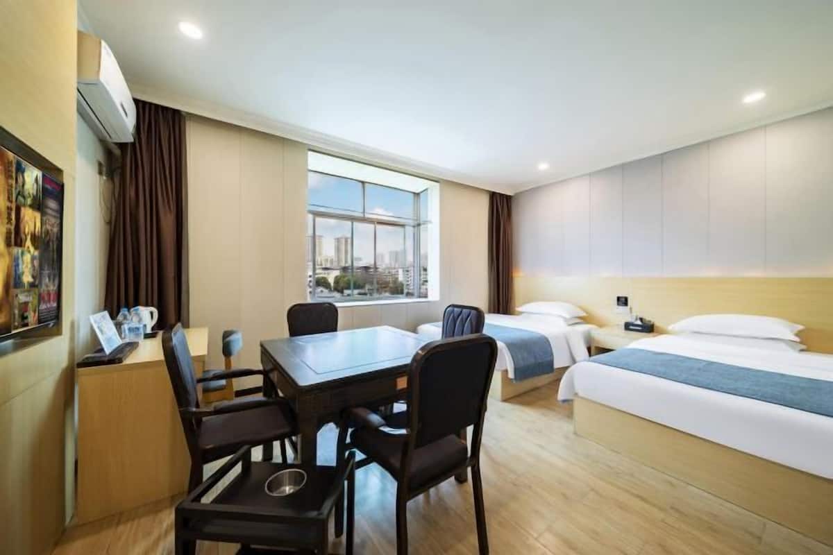 A hotel room with a bed, a desk with a chair, a window with a view of the city, and a wall-mounted air conditioner.