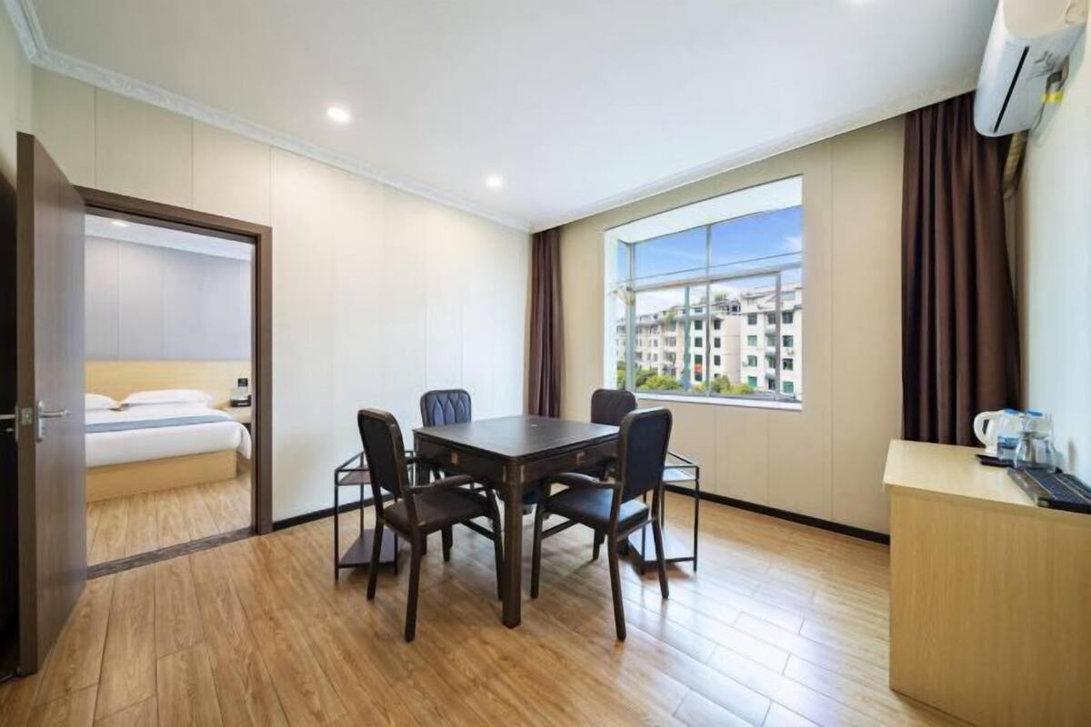 A hotel room with a wooden floor, a dining table with four chairs, a bed with white linens, a window with a city view, and a desk with a coffee maker.