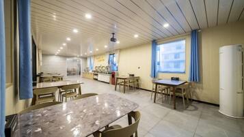 A spacious dining area with multiple tables and chairs, a kitchen area in the background, and a large window providing natural light.