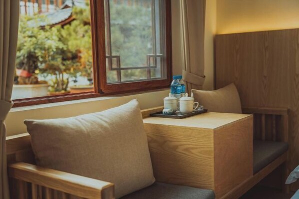 A wooden bench with cushions, a small table with a tray holding a bottle and cups, and a window with a view of greenery.