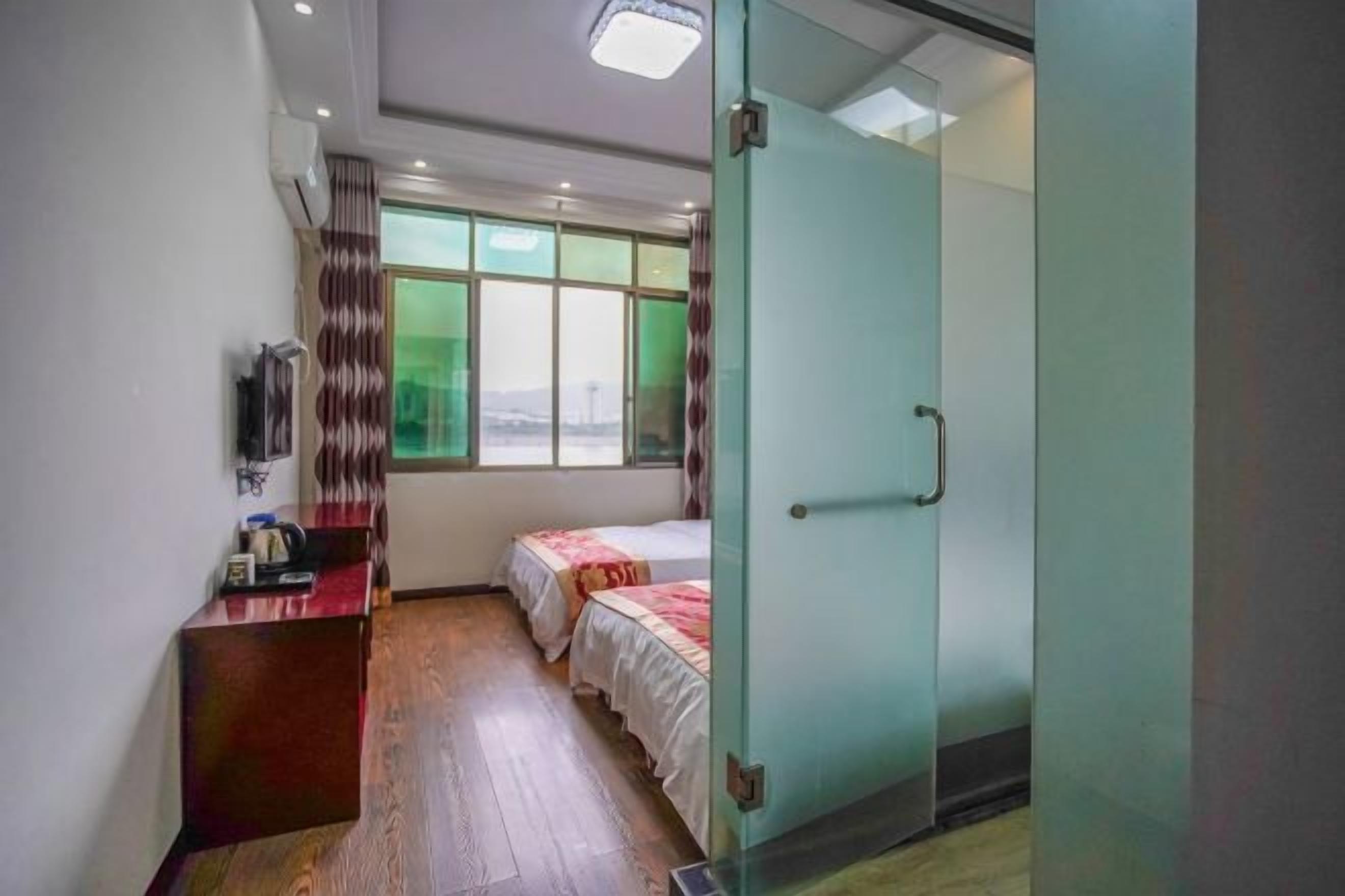 Quarto com piso de madeira, uma escrivaninha com cadeira, duas camas com roupa de cama branca, porta de vidro e janela com vista.