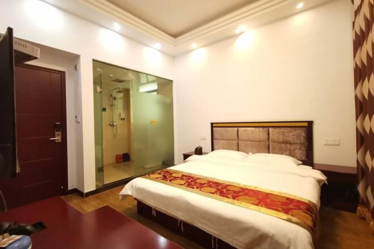 A hotel room with a bed, a wooden door, a glass shower enclosure, and a patterned curtain.