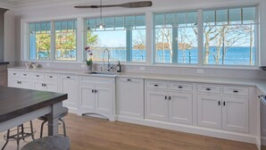 A kitchen with white cabinetry, a large window with a view of the sea, and a wooden dining table. - Waterfront estate near Portland with pool, dock and sweeping views of Casco Bay. (Yarmouth)