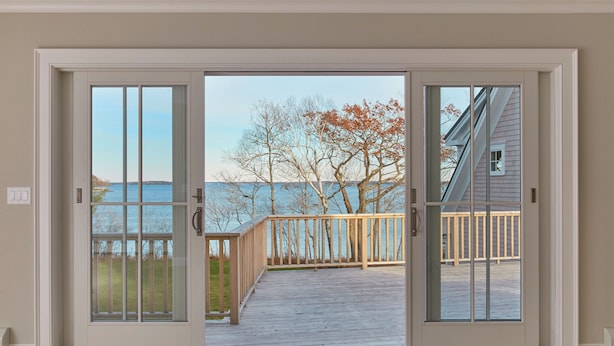 A view through a sliding glass door to an outdoor deck with a wooden railing, overlooking a body of water and trees. - Waterfront estate near Portland with pool, dock and sweeping views of Casco Bay. (Yarmouth)