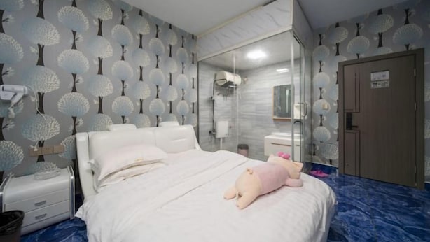 Habitación de hotel con cama, espejo, armario y baño visible tras una pared de vidrio.