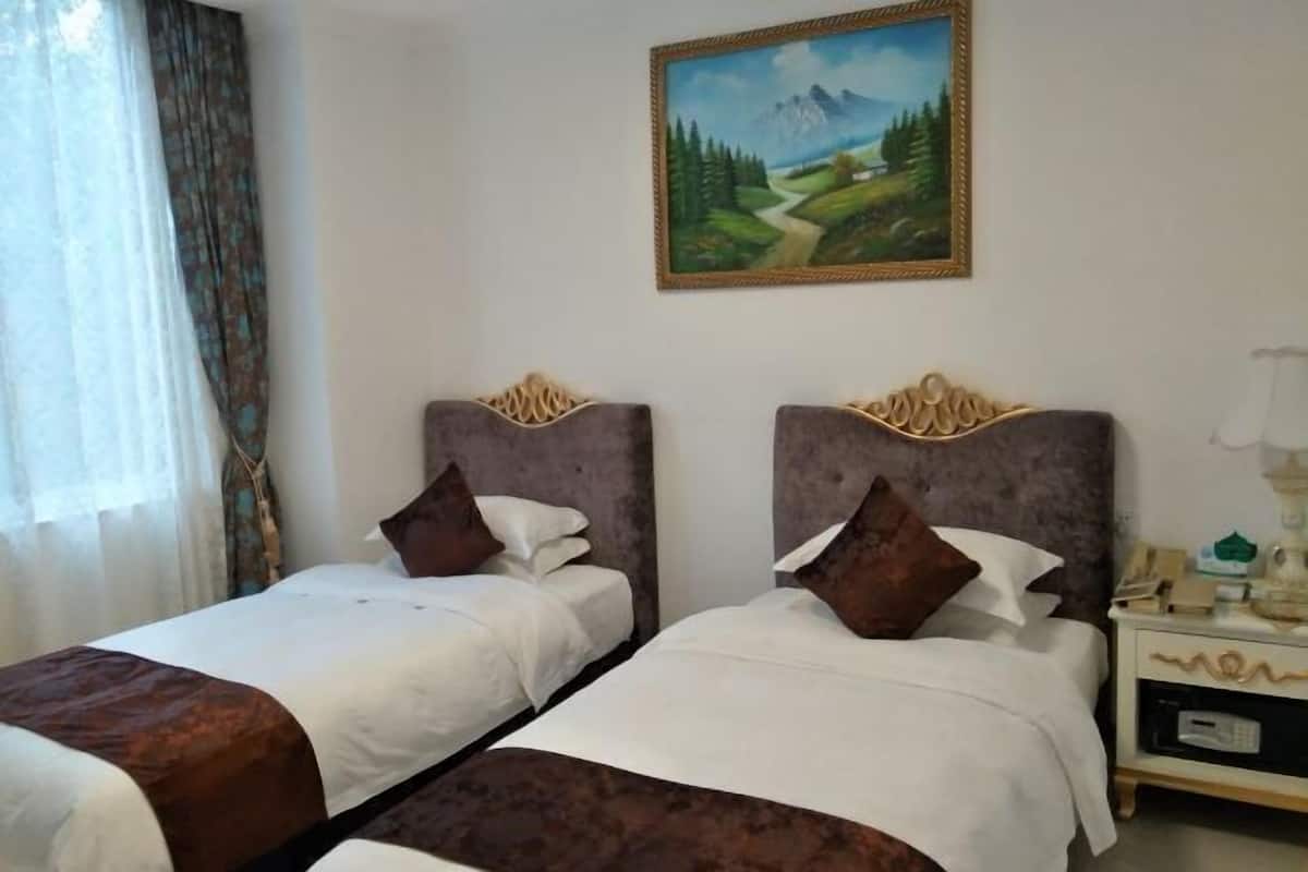 A hotel room with two beds, a painting on the wall, a nightstand with a lamp, and a window with curtains.