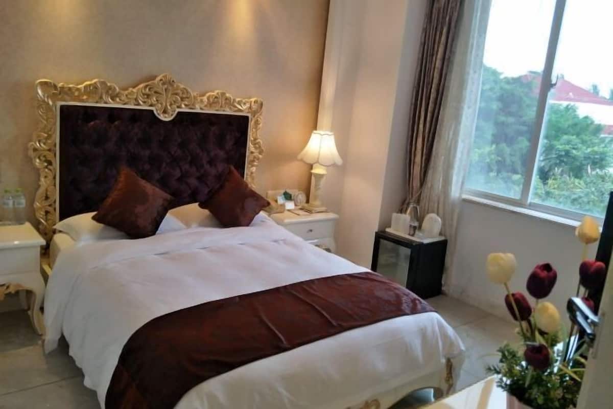 A hotel room with a large bed, a nightstand, a lamp, a window with a view, and a vase with flowers.