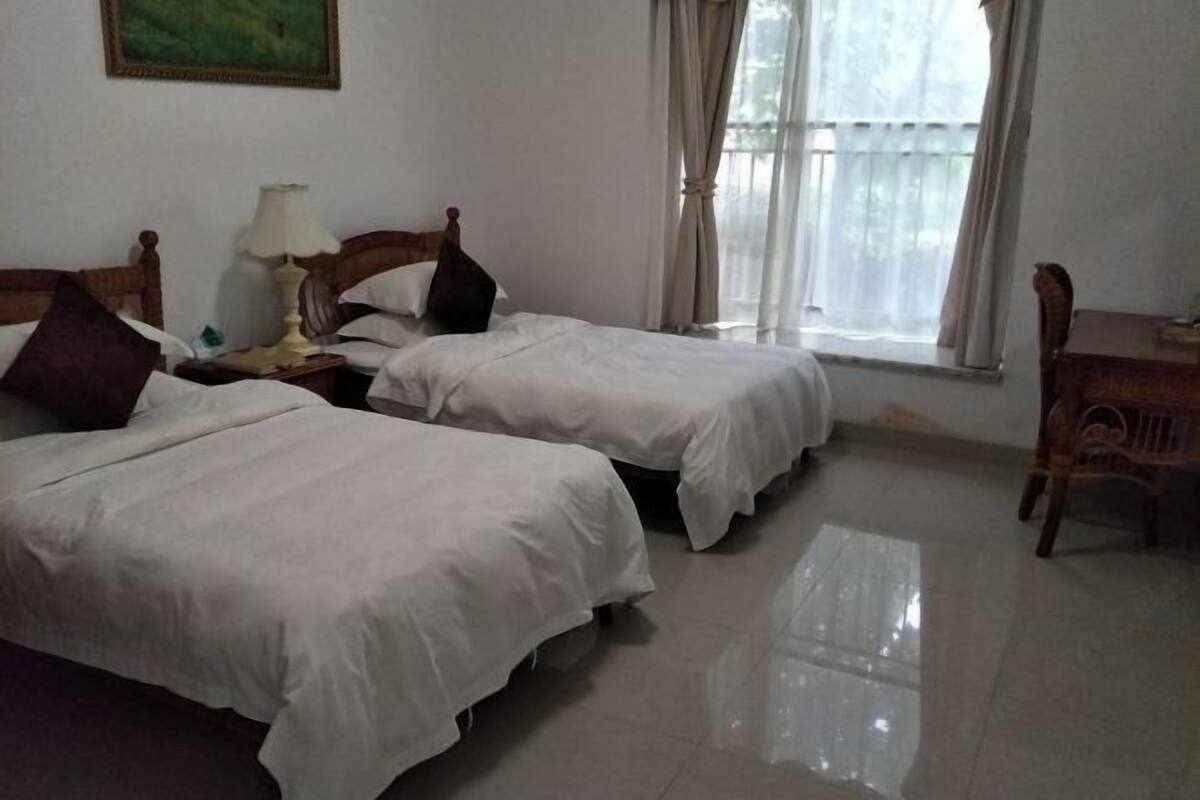 A hotel room with two beds, a nightstand, a lamp, a window with curtains, and a wooden chair.