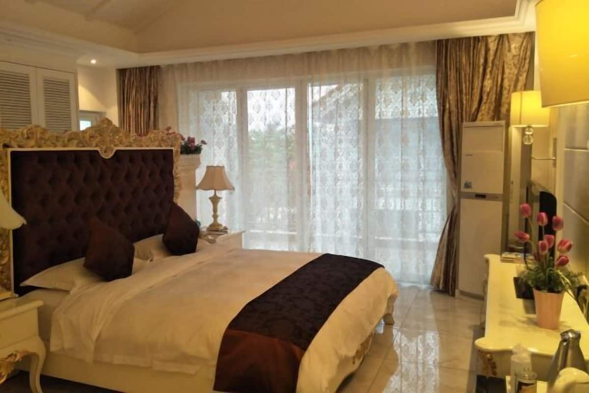 A bedroom with a large bed, a nightstand, a lamp, a dresser, and a window with curtains.