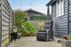 A patio with a wicker chair, a wooden deck, a potted plant, and a house with a sloped roof. - Holiday home close to the beach and restaurants (Karrebæksminde)