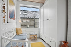 Una habitación pequeña y blanca con una cama, un taburete, un armario y una ventana con vista a una casa.