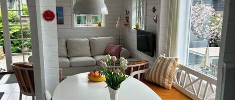 A room with a white table, a sofa with cushions, a TV, and a window with a view of greenery.