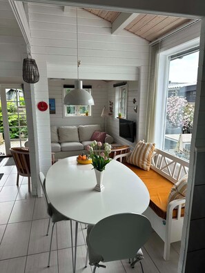 A room with a white table, a sofa with cushions, a TV, and a window with a view of greenery.