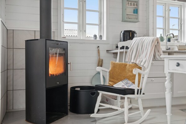 A modern kitchen with a wood-burning stove, white cabinetry, and a rocking chair with a magazine. - Holiday home close to the beach and restaurants (Karrebæksminde)
