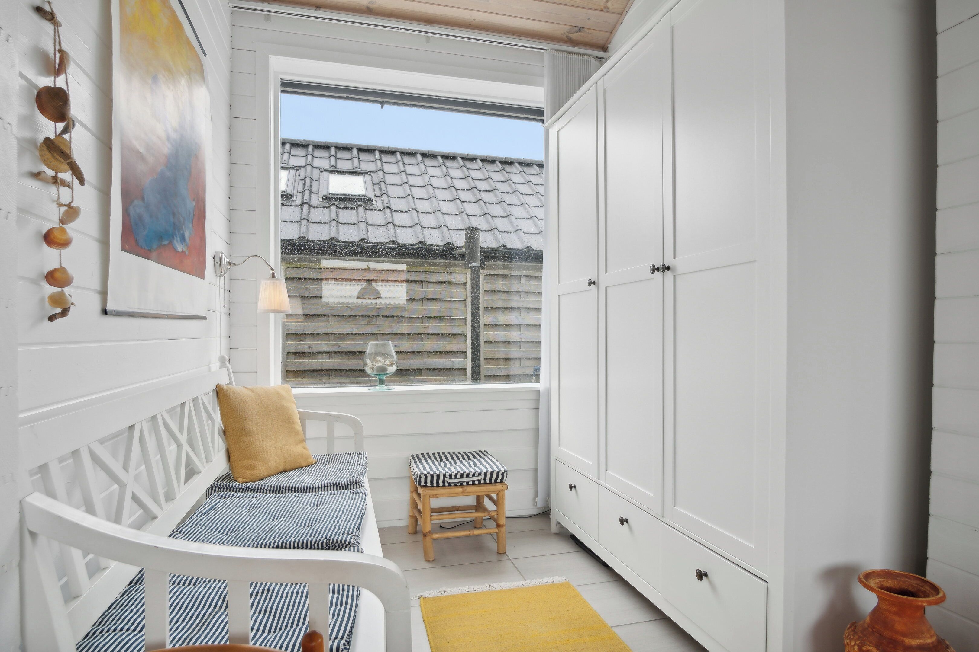 A small, white room with a bed, a stool, a cabinet, and a window with a view of a house.
