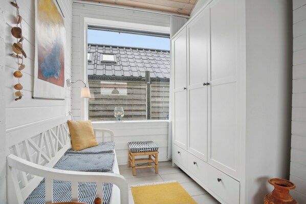 A small, white room with a bed, a stool, a cabinet, and a window with a view of a house.