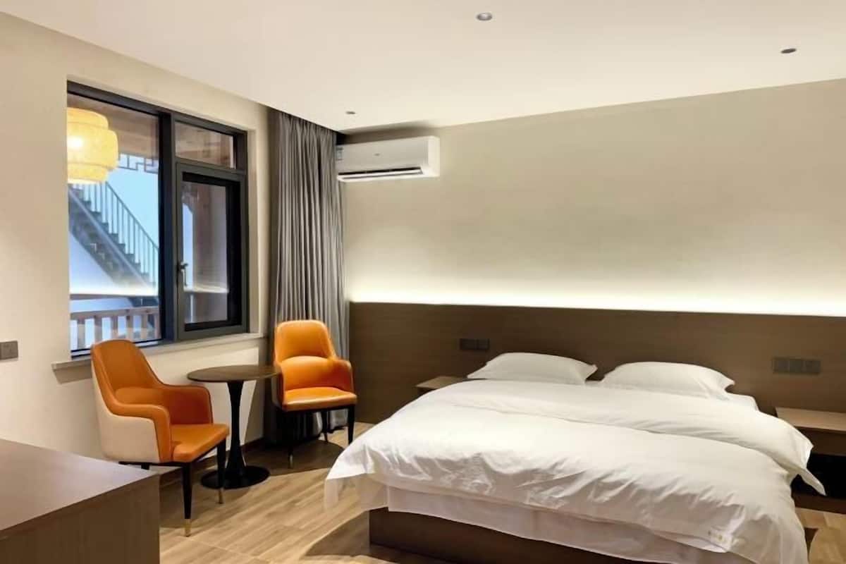 Habitación de hotel con cama, dos sillas, una mesita, aire acondicionado y ventana con vista a un edificio.