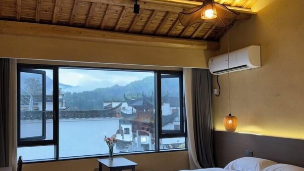 Una stanza con un letto, un tavolino con un vaso, una finestra vista edifici e montagne, e un condizionatore d'aria.