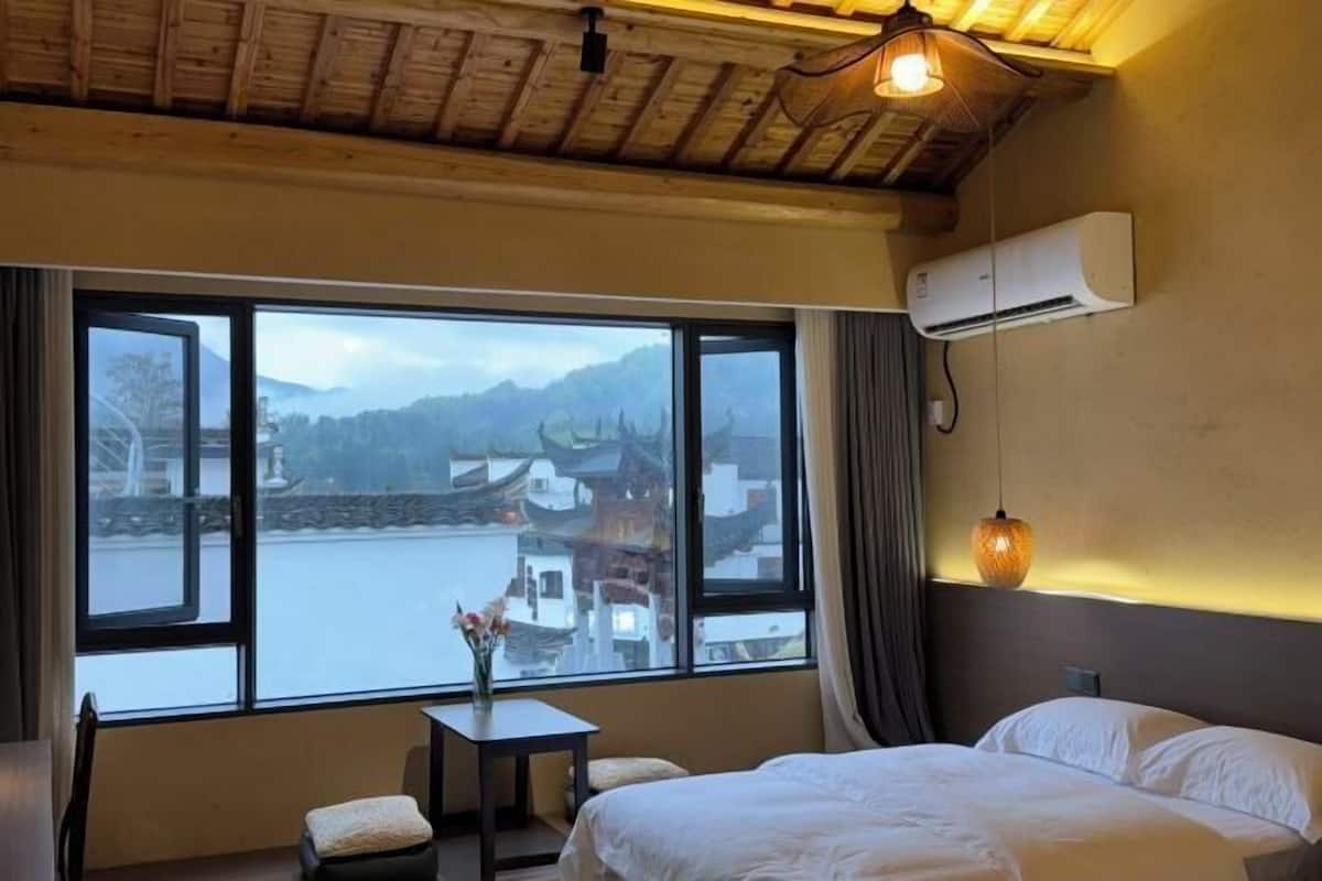 Una habitación con cama, una mesita con un florero, una ventana con vistas a edificios y montañas, y un equipo de aire acondicionado.