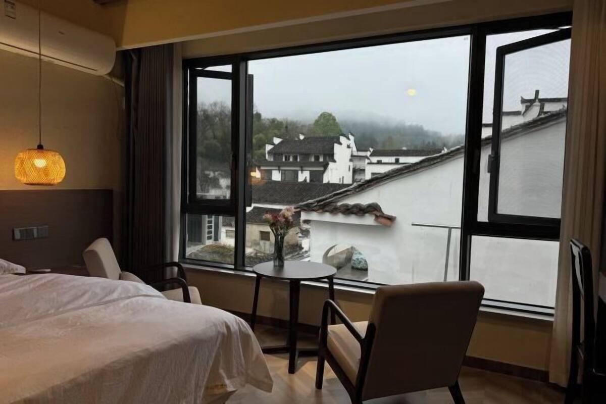 Habitación de hotel con cama, dos sillas, una mesita y un ventanal grande con vistas a edificios y un río.