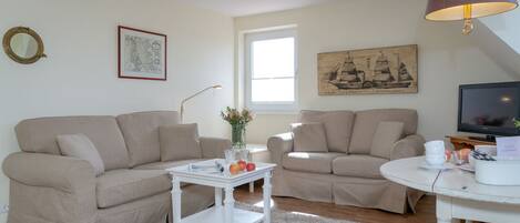 A living room with two beige sofas, a white coffee table, a small white side table, a round white table, a television, and a lamp.