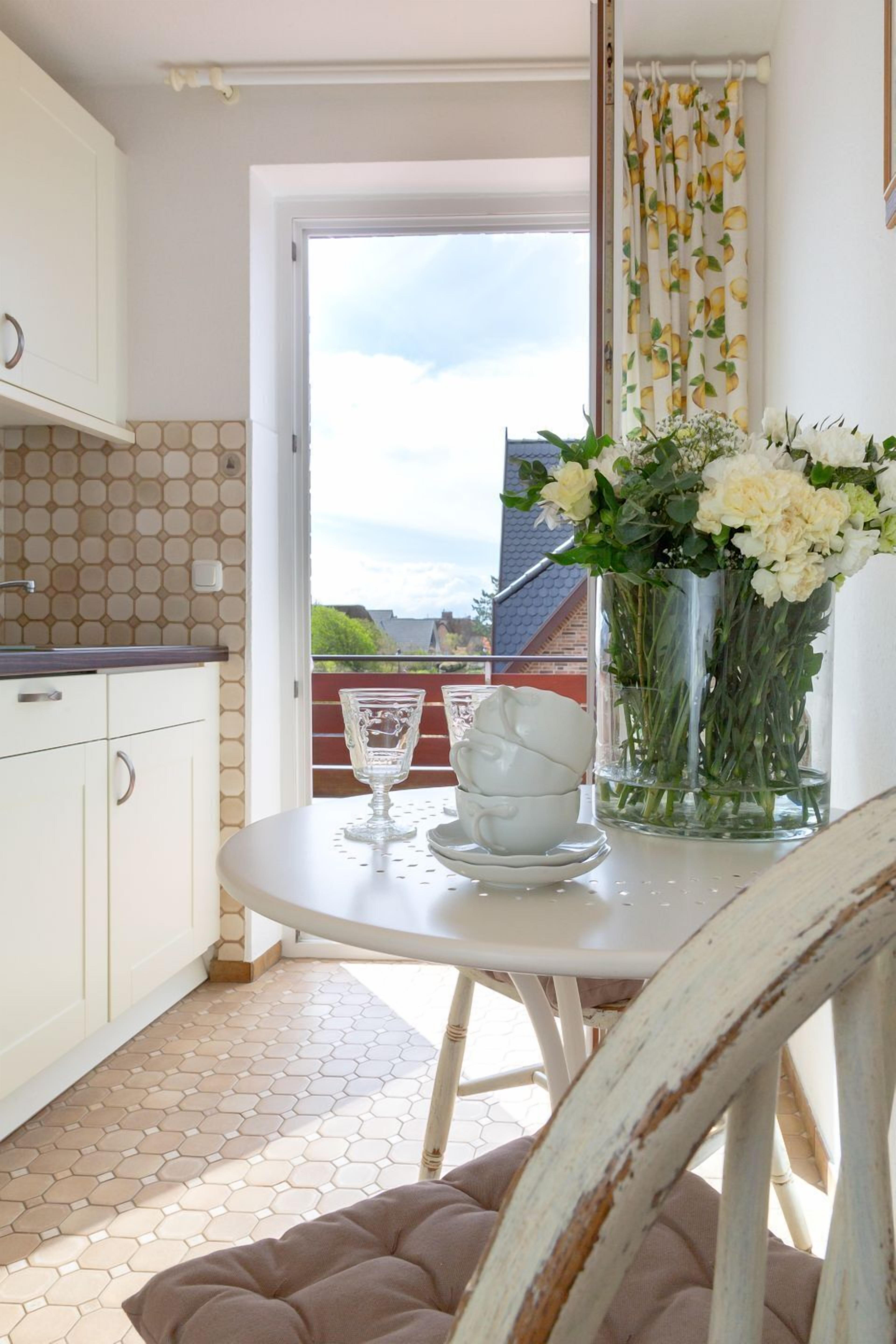 Eine Küche mit weißen Schränken, einem sechseckigen Fliesenboden, einem Esstisch mit Blumenvase und Blick durch eine Tür.