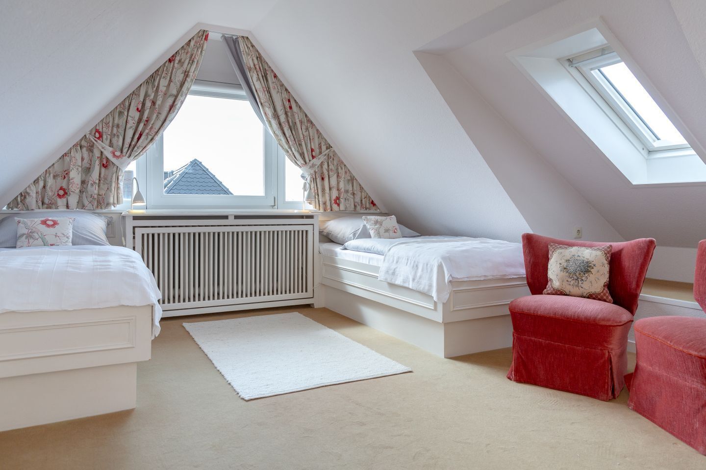Ein Schlafzimmer mit zwei Betten, einem Heizkörper, einem Fenster mit Vorhängen, einem Dachfenster und einem roten Stuhl.