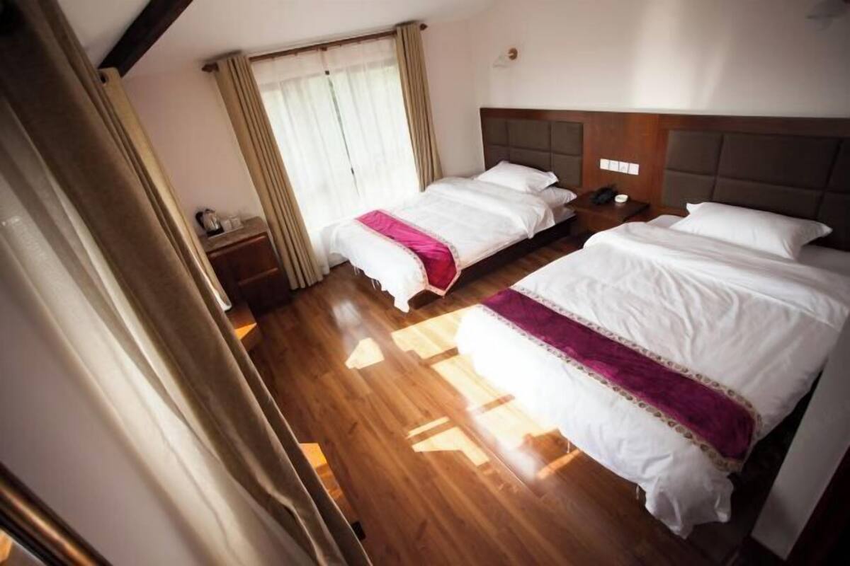 A hotel room with two beds, wooden flooring, a nightstand with a phone, and a window with curtains.