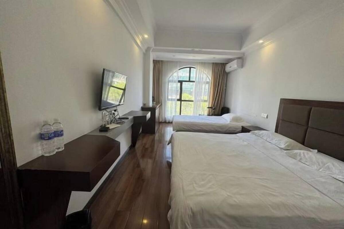 A hotel room with two beds, a desk with a flat-screen TV, a window with curtains, and a view of greenery outside.