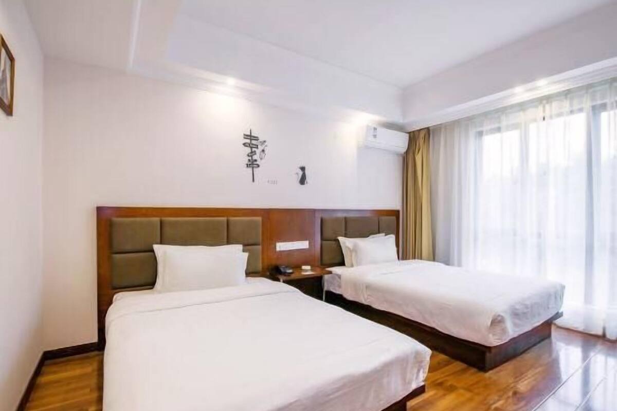 A hotel room with two beds, wooden flooring, a large window with curtains, and a wall-mounted air conditioning unit.
