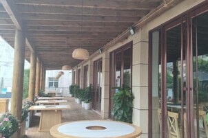 Uma área externa coberta com mesas e bancos de madeira, plantas em vasos e janelas amplas.