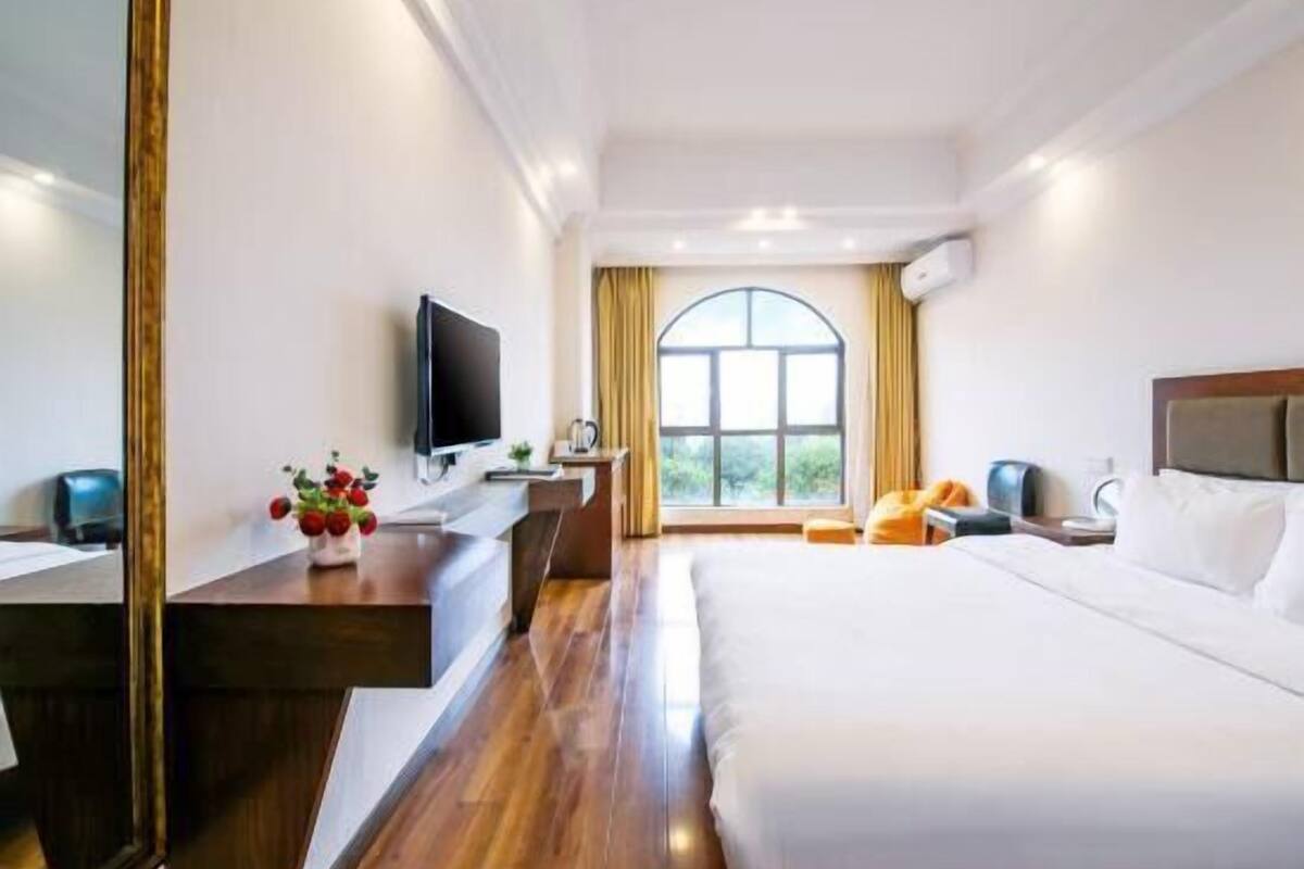 A hotel room with a bed, a desk with a flat-screen TV, a window with curtains, and a view of greenery outside.