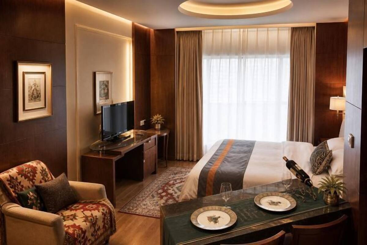 A hotel room with a bed, a desk, a chair, a dining table with plates, and a bottle.