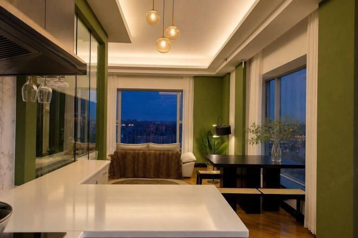 A modern hotel room with a large window, a dining area, and a kitchen.