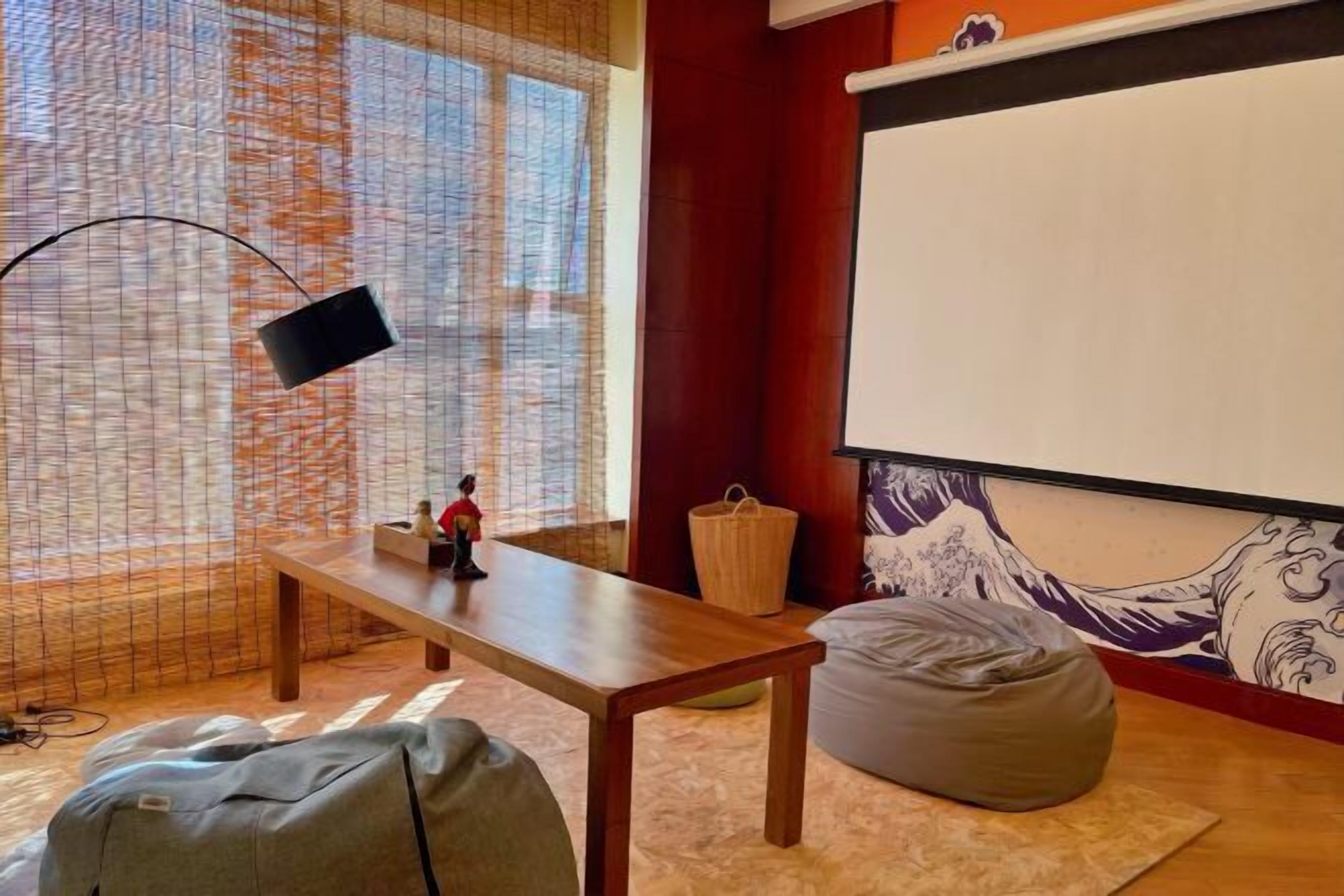 A room with a wooden table, a bean bag, a projector screen, and a large window with a view of a building.