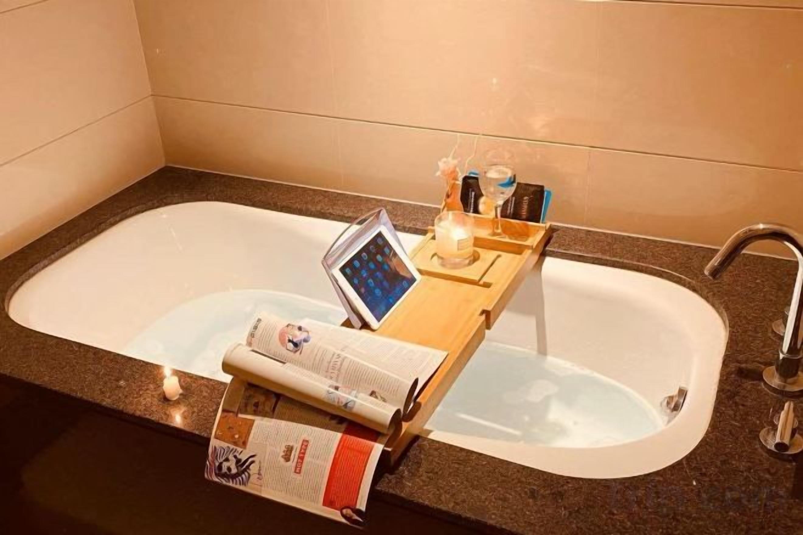 A bathroom with a bathtub, a wooden tray holding a book, a candle, and a bottle.