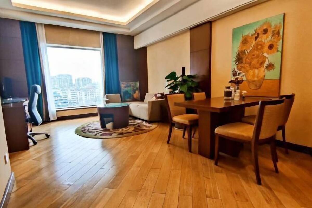 A hotel room with a dining area, a large window with a city view, a painting on the wall, and a plant on the table.