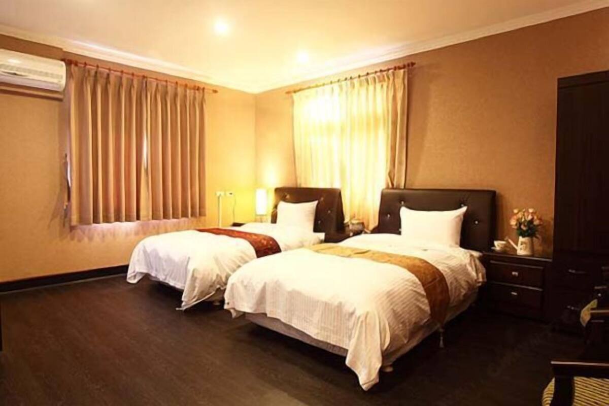 Une chambre d’hôtel avec deux lits, un plancher en bois, une fenêtre avec des rideaux, une table de chevet et une lampe.
