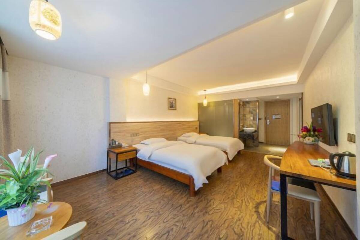 A hotel room with a wooden floor, a bed, a desk, a chair, a TV, and a plant.
