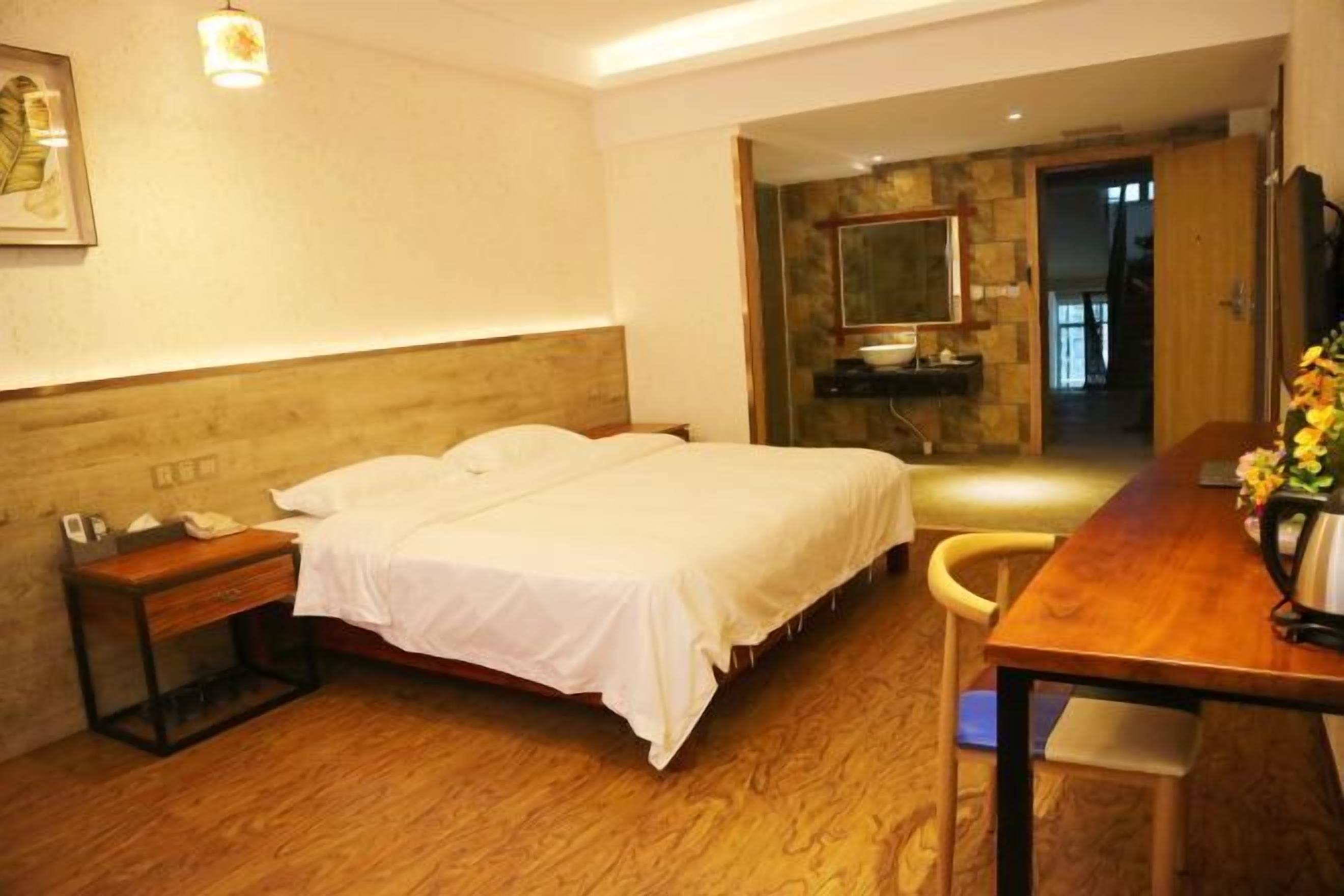Habitación de hotel con cama, mesita de noche, silla, piso de madera y un escritorio con un jarrón de flores.