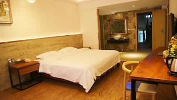 Habitación de hotel con cama, mesita de noche, silla, piso de madera y un escritorio con un jarrón de flores.